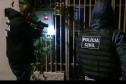 Dois policiais civis em frente a um portão. Um deles aponta uma arma.