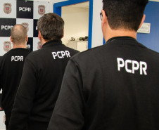 Três policiais civis de costas. Ao fundo, banner da PCPR