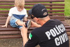 Policial civil agachado, prendendo sandália no pé de uma criança sentada em banco de madeira