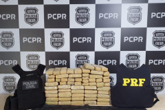 Diversos tabletes de droga entre coletes da polícia civil e policia rodoviária federal. ao fundo, banner da polícia civil