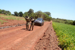Policiais andando lado a lado apontando as armas, em frente à viatura em estrada rural