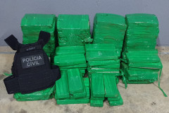Diversos tabletes de droga agrupados e um colete da polícia civil, ao lado.