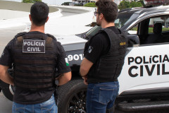 Dois policiais civis de costas, ao lado de viatura