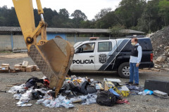 Policial civil observa retroescavadeira destruindo objetos; ao fundo, uma viatura