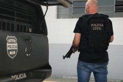 Policial civil empunhando arma, ao lado de viatura