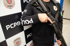 Policial civil empunhando arma em frente a um banner da PCPR