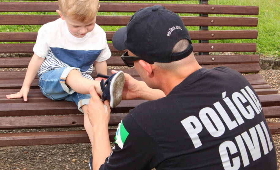 Policial civil agachado, prendendo sandália no pé de uma criança sentada em banco de madeira